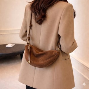 Puede incluir: Un bolso de hombro en forma de media luna de ante marrón con una correa anudada y herrajes dorados. El bolso se lleva sobre una blazer color beige, lo que sugiere un accesorio de moda.