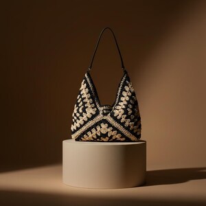 May include: A black and cream crochet bag with a black leather strap, displayed on a cylindrical plinth. The bag's crochet pattern features floral designs. The background is brown.