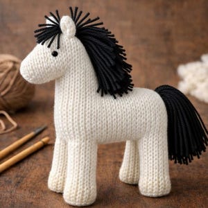 Op de afbeelding: Handgebreid wit paard met een zwarte manen en staart. Het speelgoedpaard is gemaakt met een getextureerde steek en heeft kleine zwarte kraaloogjes. Breinaalden en garen zijn zichtbaar op de achtergrond.