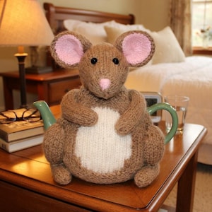 Puede incluir: Un calentador de té de ratón tejido marrón con orejas rosas y vientre blanco. El calentador está sentado sobre una tetera verde. El ratón está en una mesita de noche de madera.
