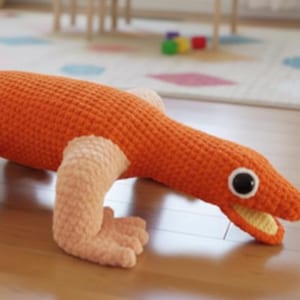 Pode incluir: Um brinquedo de crochê laranja feito à mão em forma de lagarto com pernas cor de pêssego claro e boca amarela. O lagarto tem olhos pretos e brancos e está deitado no chão de madeira. Este brinquedo macio é perfeito para crianças.