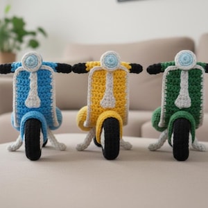 Può includere: Tre scooter all'uncinetto fatti a mano in blu, giallo e verde. Ogni scooter presenta uno pneumatico nero, un corpo bianco e grigio e manubri neri. Gli scooter sono esposti su una superficie neutra.