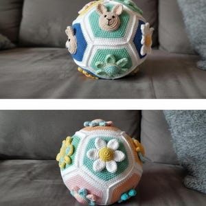 Puede incluir: Una pelota de ganchillo hecha a mano con un diseño geométrico. La pelota presenta varios colores, incluyendo blanco, azul, verde y naranja. Adornada con flores de ganchillo y caras de conejos, es un juguete suave para bebés y niños pequeños.