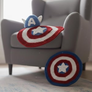 K&ouml;nnte beinhalten: Geh&auml;keltes Baby-Set im Captain America-Design, einschlie&szlig;lich einer schildf&ouml;rmigen Decke und einem passenden Hut mit Fl&uuml;geln und dem Buchstaben "A". Die Decke und der Hut sind rot, wei&szlig; und blau. Das Set wird auf einem grauen Sessel pr&auml;sentiert.
