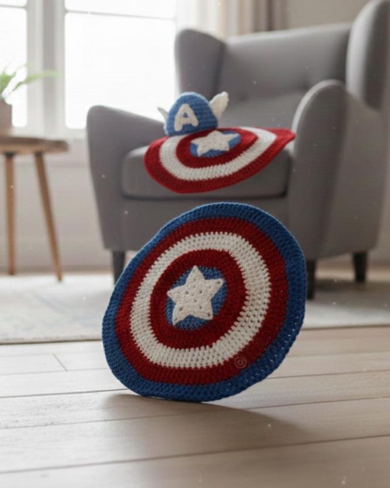 K&ouml;nnte beinhalten: Geh&auml;kelte Artikel im Captain America-Design. Das Bild zeigt einen Schild und einen Hut, beide in Rot, Wei&szlig; und Blau. Der Schild hat einen wei&szlig;en Stern, und der Hut hat ein wei&szlig;es "A" und Fl&uuml;gel. Diese handgefertigten Artikel sind f&uuml;r Kinder geeignet.