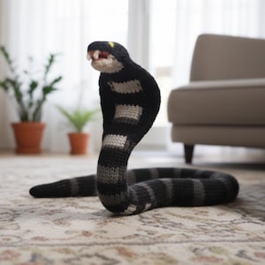 Può includere: Un giocattolo a forma di serpente cobra fatto a mano, lavorato a maglia in nero, grigio e bianco. Il serpente ha un cappuccio sollevato, bocca aperta con denti bianchi e un accento giallo sulla testa. Il corpo è a strisce e il giocattolo è posizionato su un tappeto fantasia.
