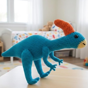 May include: A handmade crochet dinosaur toy in shades of blue, orange, and yellow. The dinosaur has a long neck, a curved orange crest, and black button eyes. It is standing on a light-colored wooden surface.