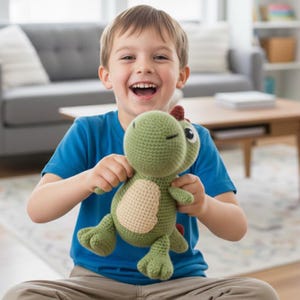 Może przedstawiać: Zielona i beżowa szydełkowa zabawka dinozaura z czerwonym grzebieniem. Dinozaur z zamkniętymi oczami jest trzymany przez uśmiechnięte dziecko w niebieskiej koszulce. Zabawka wykonana jest z miękkiej przędzy i ma szczegółowy design.