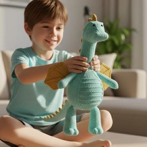 May include: A knitted teal dragon toy with yellow wings and accents. The plush dragon has a long neck, a rounded body, and a tail with yellow spikes. The toy is being held by a person wearing a light blue t-shirt.