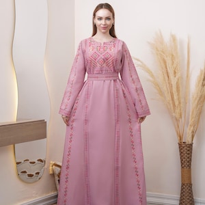 May include: A long, light pink dress with intricate embroidery. The dress features detailed floral patterns around the neckline, sleeves, and vertical panels. The long sleeves and floor-length design create an elegant silhouette. The dress has a fitted waist and a flowing skirt.