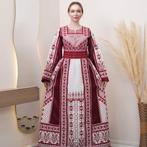 May include: A long, burgundy and white embroidered dress with long sleeves. The dress features intricate geometric patterns and a wide burgundy belt. The white skirt has detailed embroidery. The dress is worn by a person.