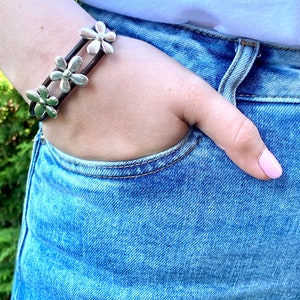 May include: A brown leather bracelet with silver flower charms. The bracelet is worn on a person's wrist.