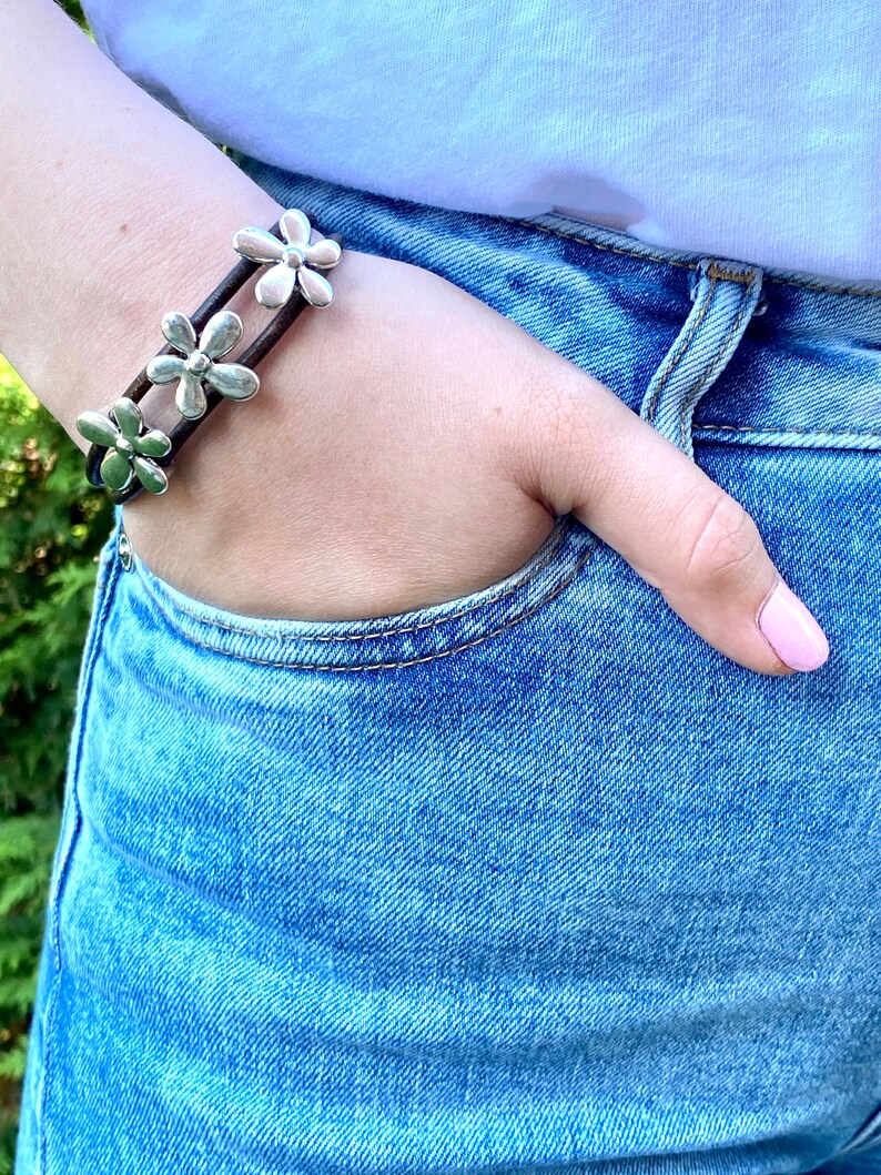 May include: A brown leather bracelet with silver flower charms. The bracelet is worn on a person's wrist.