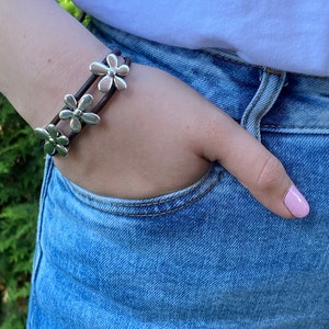 May include: A brown leather bracelet with silver flower charms. The bracelet is worn on a person's wrist.