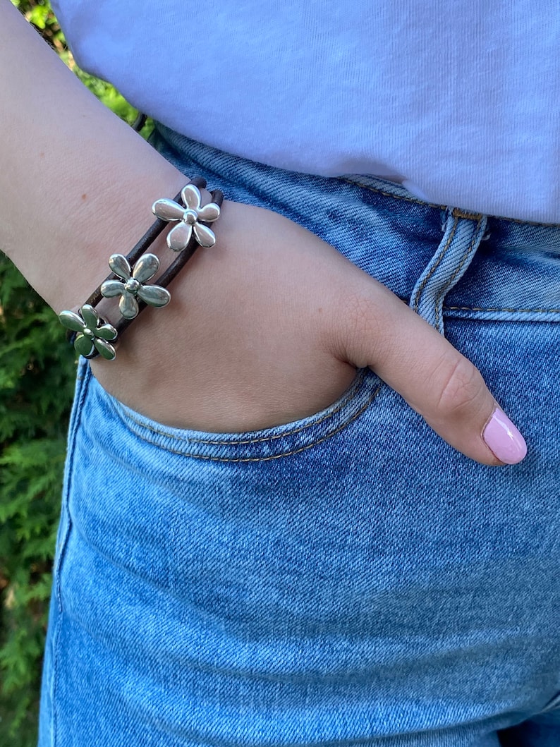 May include: A brown leather bracelet with silver flower charms. The bracelet is worn on a person's wrist.