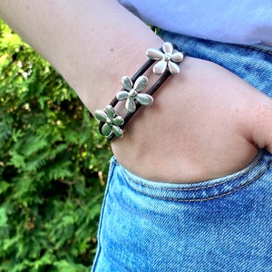 May include: A brown leather bracelet with three silver flower charms. The bracelet is worn on a person's wrist.