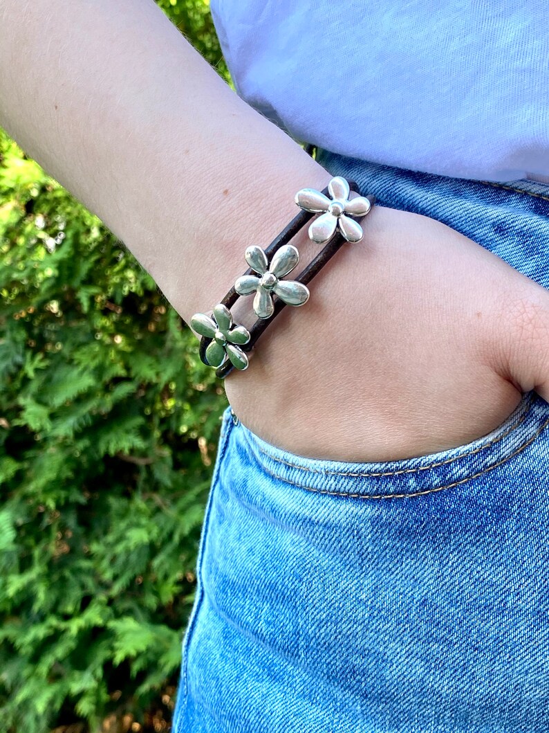 May include: A brown leather bracelet with three silver flower charms. The bracelet is worn on a person's wrist.