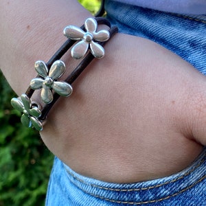 May include: A brown leather bracelet with three silver flower charms. The bracelet is worn on a person's wrist.