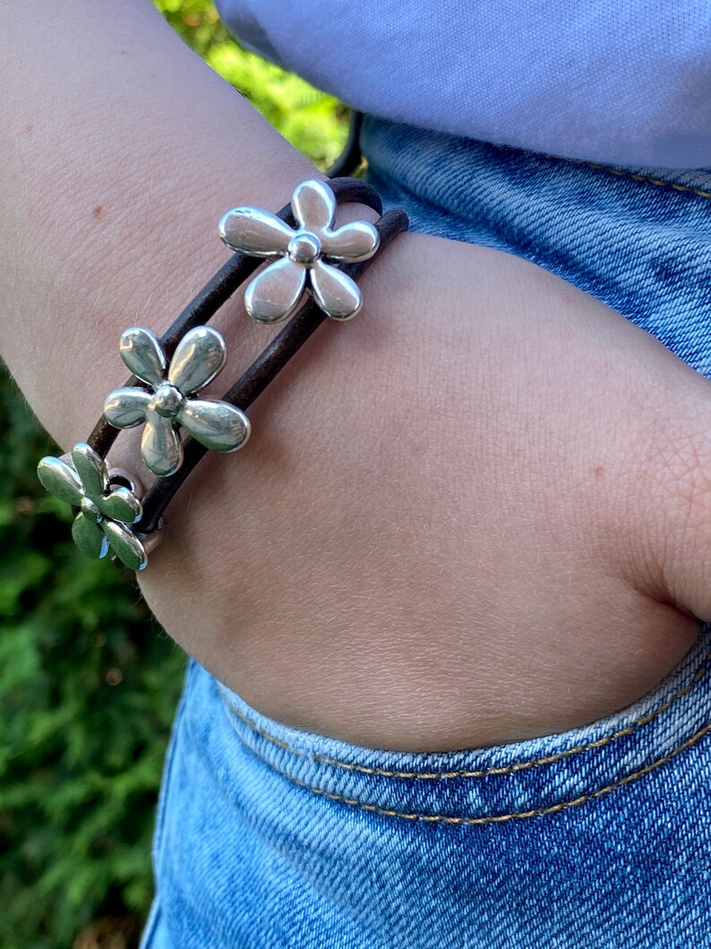 May include: A brown leather bracelet with three silver flower charms. The bracelet is worn on a person's wrist.
