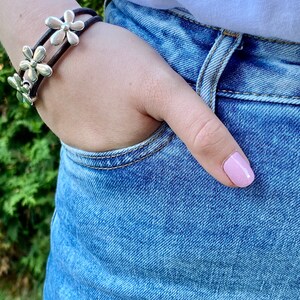May include: A brown leather bracelet with silver flower charms. The bracelet is worn on a person's wrist.