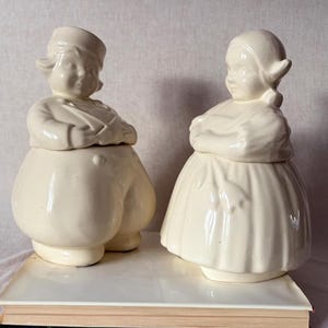 May include: Two off-white ceramic cookie jars shaped like Dutch children. The boy jar wears a hat and puffy pants, while the girl jar wears a bonnet and skirt. Both have their arms crossed and are sitting on a book.