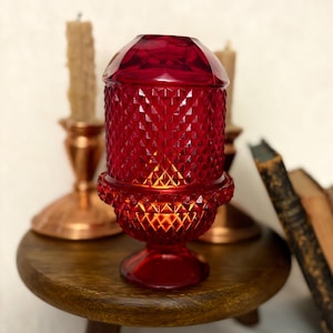 Könnte beinhalten: Ein rubinroter Glas-Kerzenhalter mit Rautenmuster, der auf einem kleinen Holztisch steht. Der Kerzenhalter hat einen Deckel und einen Sockel. Weitere Kerzen und kupferne Kerzenhalter befinden sich im Hintergrund.