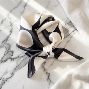 May include: A square scarf featuring a bold, high-contrast pattern of large, rounded black shapes against a smooth, off-white background. The fabric has a subtle sheen and is tied into a loose knot, creating soft folds that highlight the graphic design.