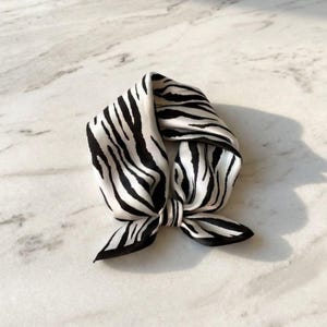 Könnte beinhalten: Ein quadratisches Seidentuch mit einem Zebra-Print in Schwarz und Weiß. Das Tuch ist in der Mitte geknotet, wobei die Enden nach außen zeigen. Das Tuch besteht aus einem weichen, fließenden Material.