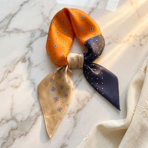 May include: A square silk scarf featuring a color-blocked design in orange, navy blue, and beige. The orange section has a small diamond pattern, the navy blue section features white polka dots, and the beige section displays a repeating square motif.