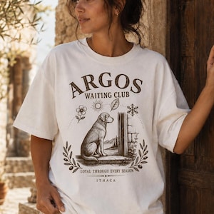 May include: An off-white short-sleeve t-shirt featuring a brown graphic print of a dog sitting by a doorway. The text reads ARGOS WAITING CLUB, LOYAL THROUGH EVERY SEASON, and ITHACA, accompanied by illustrations of a flower, sun, leaf, and snowflake.