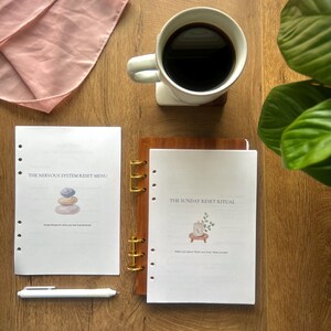 Puede incluir: Vista superior de una superficie de madera con una taza de café blanca, una bufanda rosa y dos cuadernos abiertos. Los cuadernos tienen los títulos "The Nervous System Reset Menu" y "The Sunday Reset Ritual". También se ve un bolígrafo blanco.