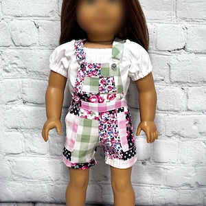 May include: A doll with long brown hair wearing a white short-sleeved shirt and patchwork overalls featuring pink floral, green gingham, and black polka dot patterns. The doll is also wearing pink and white lace-up sneakers with white rubber soles.