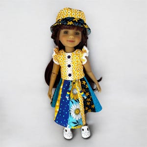 May include: A doll wearing a yellow floral print outfit and matching hat. The outfit includes a yellow top with white lace trim and blue buttons, and a patchwork skirt with blue, yellow, and floral patterns. White shoes complete the look.