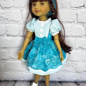 May include: A doll wearing a teal dress with a white lace trim and a matching bow. The doll has brown hair and is wearing a teal headband. The doll is also wearing white shoes. The dress has a floral pattern.