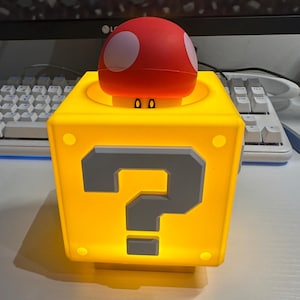 Może przedstawiać: Ż&oacute;łta, podświetlana kostka z dużym szarym znakiem zapytania z przodu. Na g&oacute;rze czerwony grzyb z białymi kropkami. Prawdopodobnie lampa lub element dekoracyjny.