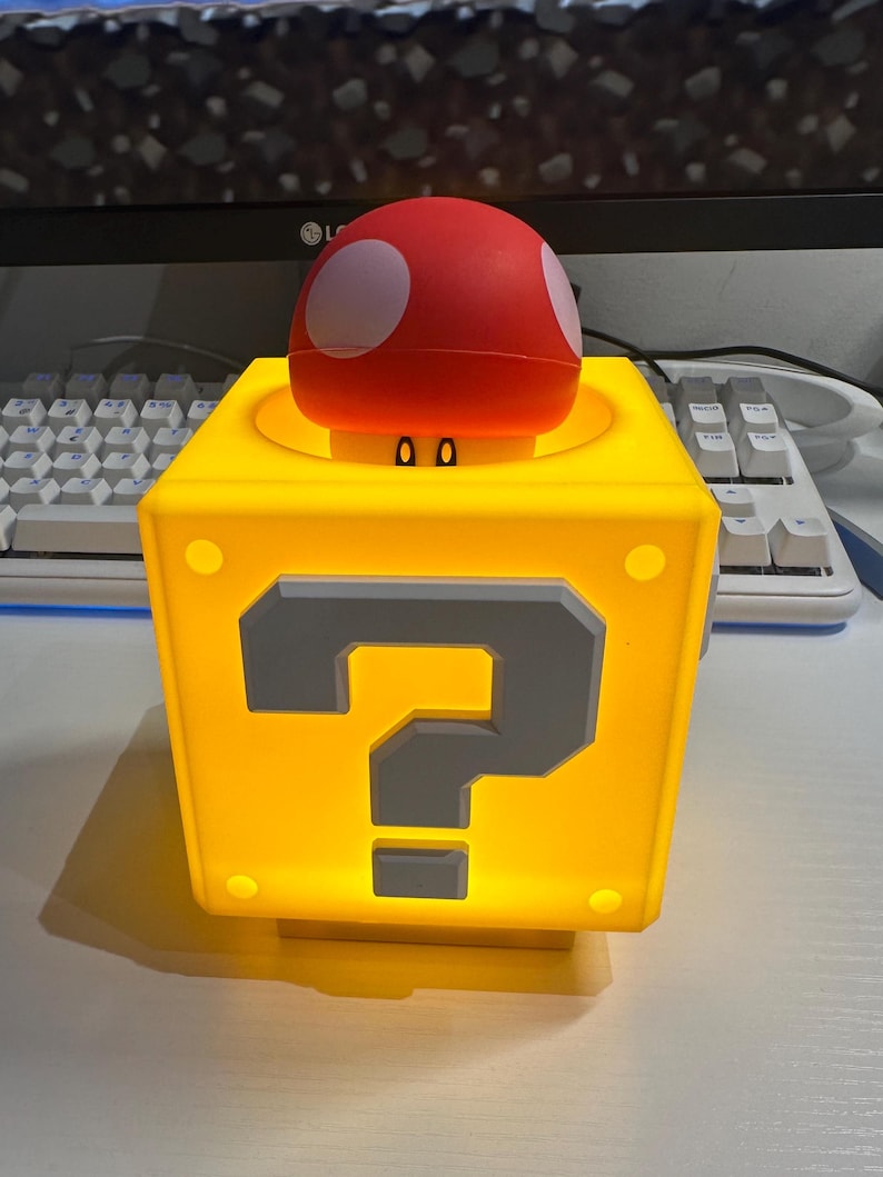 Może przedstawiać: Ż&oacute;łta, podświetlana kostka z dużym szarym znakiem zapytania z przodu. Na g&oacute;rze czerwony grzyb z białymi kropkami. Prawdopodobnie lampa lub element dekoracyjny.