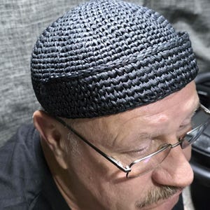 Puede incluir: Un gorro redondo de color gris carbón oscuro sin ala, hecho de un material texturizado similar a la paja, tejido apretadamente, con un detalle de borde enrollado distintivo alrededor de la base, creando una silueta estructurada.
