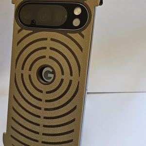 May include: Tan phone case with a circular design and the Google logo. The case has cutouts for the camera and flash. The case is made of a textured material and has a black border.