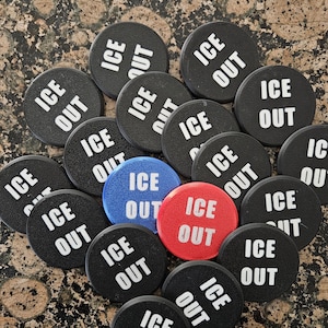 May include: A collection of black, circular tokens with the words "ICE OUT" printed in white. Two tokens are blue and red, also with "ICE OUT" in white. The tokens are arranged on a speckled brown surface.