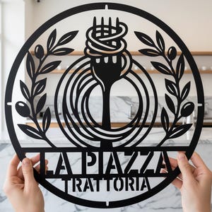 May include: Black metal wall art featuring a fork with spaghetti, olive branches, and the words "LA PIAZZA TRATTORIA" in a circular design. The artwork is held up by hands.