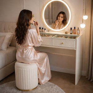May include: A white vanity table with a round, illuminated mirror. The vanity has two drawers and is accessorized with perfume bottles and makeup. A person in a pink robe sits at the vanity, applying makeup. A cream-colored, round stool with a gold base is in front of the vanity.