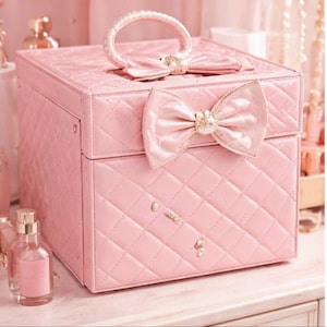 Puede incluir: Una caja de joyería rosa, acolchada y cuadrada, con un lazo en la parte delantera y superior. La caja tiene un asa y está decorada con pequeños adornos decorativos. La caja está sobre una superficie blanca.