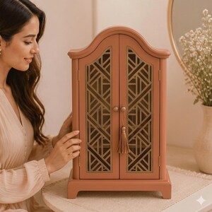 May include: A coral-colored jewelry armoire with gold geometric lattice design on the doors. The armoire has a decorative tassel and is set on a table. A woman is seen in the background.