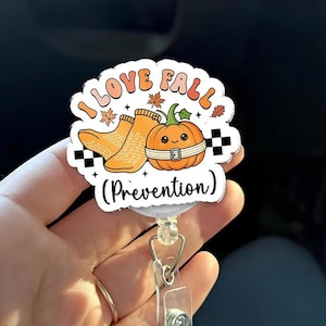 May include: A circular badge reel featuring a cartoon orange pumpkin with a face and a white belt, yellow socks, and autumn leaves. The text reads I LOVE FALL (Prevention) in black and orange letters with a black and white checkered border design.