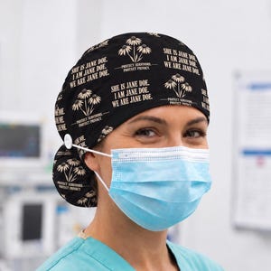 May include: A black scrub cap featuring a repeating pattern of white flowers and the text "SHE IS JANE DOE. I AM JANE DOE. WE ARE JANE DOE." along with "PROTECT SURVIVORS. PROTECT PRIVACY." The cap includes a white button on the side for mask attachment.