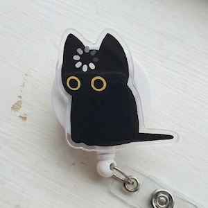 Op de afbeelding: Een witte intrekbare badgehouder met een illustratie van een zwarte kat met grote gele ogen en een cirkelvormig laadicoon op het voorhoofd. De kat is gedrukt op een doorzichtige acryllaag die is bevestigd aan een ronde witte plastic behuizing.