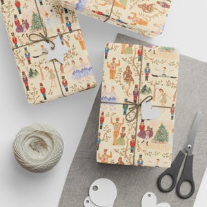 Op de afbeelding: Crèmekleurig inpakpapier met een herhalend patroon van notenkrakers, ballerina's en kerstbomen, vastgebonden met donkerbruin touw en witte ster- of hartvormige cadeaulabels. Inclusief een bol touw en een zwarte schaar.