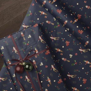 Op de afbeelding: Donkerblauw inpakpapier met een herhalend patroon van notenkrakers, kleine cadeaudoosjes, hulsttakjes en gouden sterren. Een rechthoekig cadeau verpakt in dit papier is gestrikt met een donkerrood lint en twee ronde kerstballen.