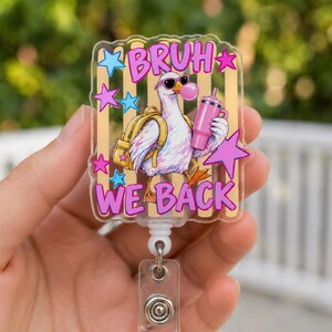 May include: A clear acrylic badge reel featuring a cartoon duck wearing sunglasses and a yellow backpack while holding a pink tumbler. The duck is blowing a pink bubble. The text BRUH WE BACK is printed in pink, surrounded by blue and pink stars.