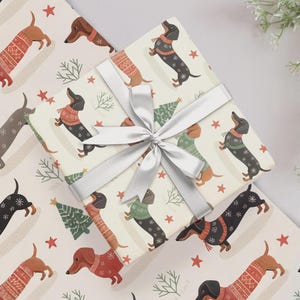 May include: A square gift box wrapped in cream-coloured paper featuring a repeating pattern of dachshunds wearing jumpers, green pine trees, and red stars. The package is tied with a wide, smooth, silver satin ribbon in a centred bow.